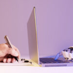 Human hand writing on a notepad with a pencil opposite a robot hand typing on a laptop keyboard